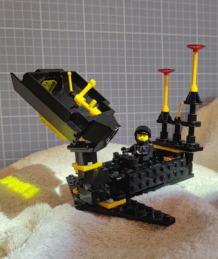 A small Lego Blacktron build, with an open antenna dish at the front, and a smiling minifig sitting inside the cockpit. The build is mostly in black bricks, with a few yellow accents.
