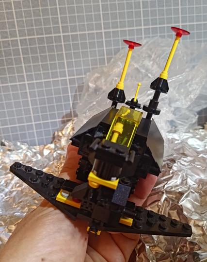 A Lego Blacktron build of a small spaceship. The build is mostly in black bricks, with a few yellow accents. A hand is holding it above a simple background diorama mimicking a metallic asteroid surface.