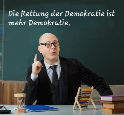 Lehrer sitzt hinter seinem Schreibtisch, erhobener Zeigefinger, an der Tafel hinter ihm spruch:
Die Rettung der Demokratie ist mehr Demokratie.