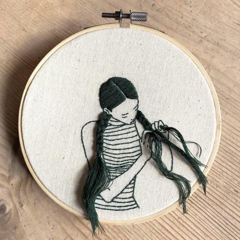 Photograph featuring an embroidery hoop on a pale wooden surface with an image of a figure in black thread sewn on tbe cream material the figure a young woman is shown braiding her long hair with thread that produdes from the image and wears a striped top