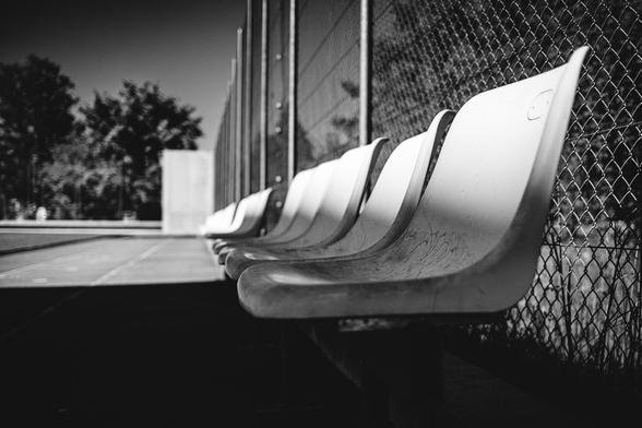 Black and white photo of empty plastic stadium seats lined up against a chain-link fence, with strong shadows and sunlight creating dramatic contrast. The scene appears quiet and still, likely beside a sports court or field.
