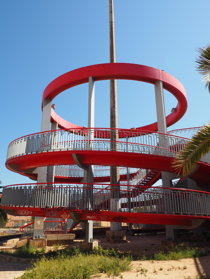 Hochkantfoto: Eine spiralförmige Fahrradauffahrt mit zwei Umdrehungen führt auf eine Brücke. Oben drüber hängt noch ein Kreis der die Träger stabilisiert, in der Mitte steht ein hoher Mast mit Beleuchtung die nicht mit im Bild ist.