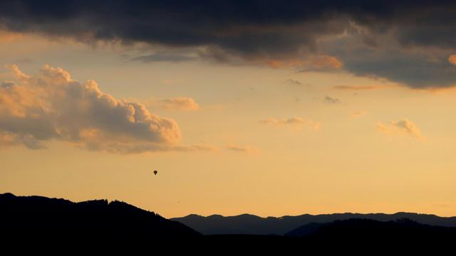 Evening photo looking at a hilly landscape that is already dark. The sky is yellow/orange from the light of the setting sun. Large heavy dark clouds are in the top of the frame. There is an hot air balloon back in the distance visible only as a small black silhouette.
