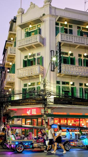 color photo in portrait format of a street corner of Chinatown taken after sunset with an early 20th century 3 stories western style residential build in cream off-white color and a restaurant in red neon lights occupying the ground floor. several tuktoks parked in front.