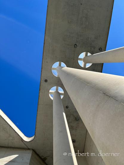 Drei runde Betonsäulen ragen von rechts unten in die Mitte des Bildes hinauf gen Himmel, hoch hinauf, in eine offene Lochstützenkonstruktion, die das gewaltige Vordach trägt, vor einem tieflbauen Himmel.