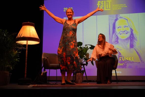 Cornelia Funke in einem blumigen Sommerkleid ohne Ärmel mit weit ausgebreiteten Armen vor ihrem Stuhl auf der Bühne. Rechts dahinten auf dem Stuhl Jagoda Marinić. Im Hintergrund auf einer Leinwand auch ein Bild von Funke. Und links eine alte Stehlampe.