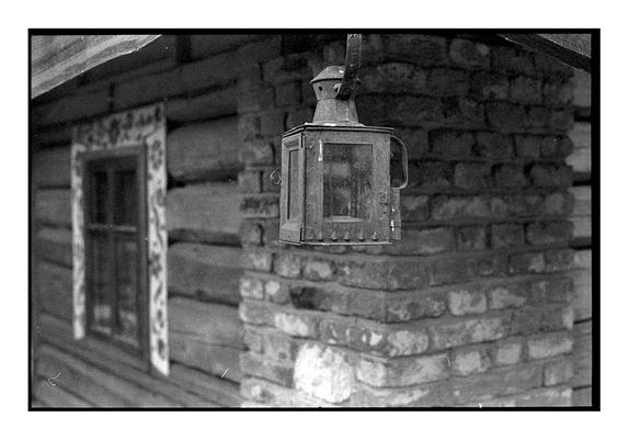 A close-up of an old metal lantern hanging outside a rustic wooden and brick house. The background shows a window with traditional floral decoration. Captured on Ilford Delta 3200 black and white film.
