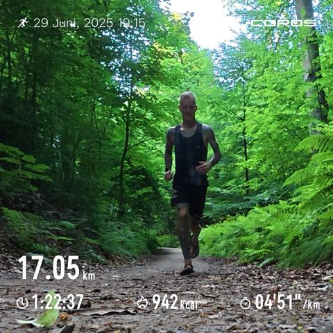 Das Bild zeigt einen Mann, der auf einem Waldweg joggt. Der Weg ist von dichtem, grünen Laub umgeben.  Im Bild sind auch Daten eines Fitness-Trackers zu sehen, die die gelaufene Distanz (17,05 km), die Laufzeit (1:22:37), den Kalorienverbrauch (942 kcal) und das Tempo (4 Minuten 51 Sekunden pro Kilometer) angeben.  Der Mann trägt ein ärmelloses Shirt und Sandalen. Das Bild wirkt insgesamt ruhig und naturverbunden.