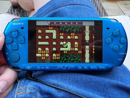 A photo of my blue PSP resting on my lap and showing the Mega Drive game Mega Bomberman. This area has some brick walls, trees at the top and bottom and a river with a small rope bridge across it to the right.