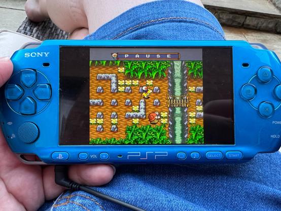 A photo of my blue PSP resting on my lap and showing the PC Engine game Bomberman ‘94. This area has some brick walls, trees at the top and bottom and a river with a small rope bridge across it to the right. It is the same as in yhr previous photo, however this version has much more vibrant colours than in the Mega Drive version.