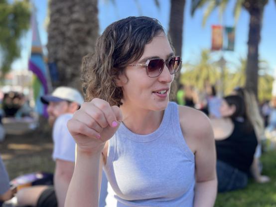 A beautiful woman with curly brown hair wearing sunglasses and a gray tank top. She is sitting in the park with a half smile on her half-parted lips as she explains something to someone off-camera on the right. The smile brings out the dimple in her check, and the beautiful shape of her cheekbones. Her right hand is raised as if emoting, showing pink nail polish. In the blurred background, palm trees, other people, and a pride progress flag.