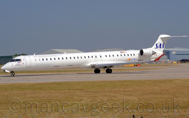 Side view of a long, thin, twin engined jet airliner taxiing from right to left as it turns on to a grey runway.
The plane is mostly white, with grey "Scandinavian" titles on the upper forward fuselage, with additional "Airlines" titles on the lower forward fuselage.
Smaller "Scandinavian" titles are on the sides of the white engine pods, mounted on the sides of the rear fuselage, just in front of and below the tail.
The blue registration "EC-MLC" is on the lower rear fuselage, just aft of a small Spanish flag, which itself is just aft of a stylised golden crown.
On the upper rear fuselage, there is a 9*3 grid of coloured dots, representing the flags of Norway, Sweden, and Denmark, the 3 countries for whom SAS is the nominal flag carrier.
Slightly yellowing grass fills the foreground, along the side of the runway, while grey hangars are visible over the top of the plane in the background, under blue skies.