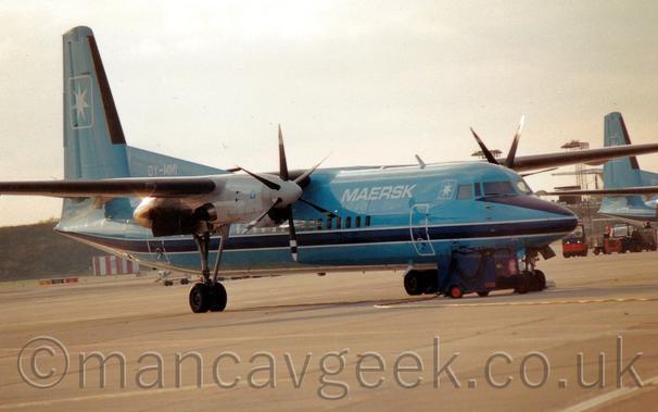 Side view of a high-winged, twin propellor-engined airliner parked facing to the right.
The plane is a light blue, with a dark blue stripe outlined in white running along the body under the passenger cabin windows, and white "Maersk" titles on the upper forward fuselage.The white registration "OY-MMI" is on a fillet at the front of the tail.
There is the white outline of a square containing a 7-pointed star on the tail, repeated in smaller form just aft of the cockpit windows.grey concrete apron fills the foreground, with a grass berm in the background coming in from the left.The tail of a similar plane can be seen on the right of the frame.
Bright but hazy skies fill the rest of the frame.