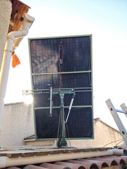 Solar panels mounted on a metal frame, which is itself fixed to a roof between two houses. A system of hydraulic jacks is visible, suggesting the mobility of the whole.