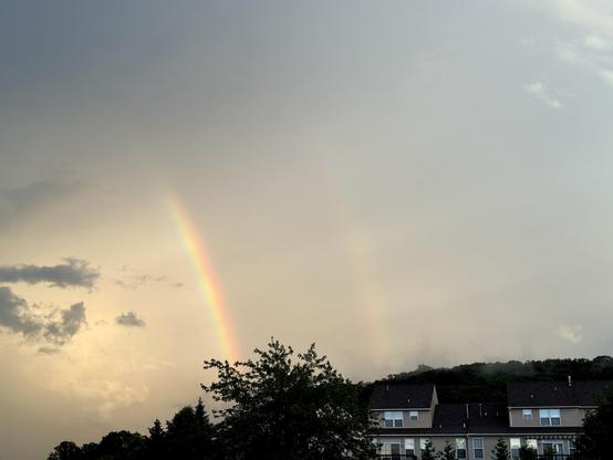 Gray, cloudy sky with a double rainbow over some trees and houses.