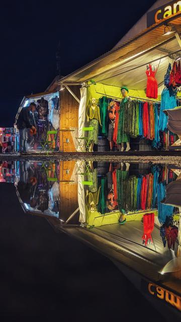 Das Bild zeigt einen farbenfrohen Marktstand bei Nacht, der Kleidung verkauft. Der Stand ist intensiv beleuchtet und hebt sich leuchtend vom dunklen Hintergrund ab. Ein auffälliges rotes Kleid hängt im Zentrum, umgeben von weiteren bunten Kleidungsstücken, die ordentlich präsentiert sind. Unter dem Stand befindet sich eine Pfütze, die das gesamte Szenario klar reflektiert. Diese Spiegelung verdoppelt die Farben und Formen, wodurch eine faszinierende Symmetrie entsteht. Vor dem Stand stehen zwei grüne Stühle und ein ebenso grüner runder Tisch, und eine Person betrachtet die Ware.