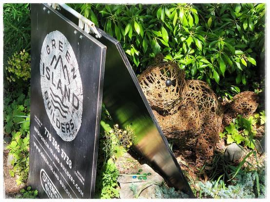 This image shows a black and white sandwich board advertising Green Island Builders. Their logo shows the cut end of a log. The sign is perched at the end of someone's lawn. Immediately behind the board is a large lawn ornament of a wire mesh beaver. An appropriate mascot?
