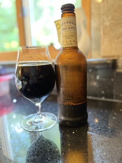Highlighted by sunlight from a window in the background, a tall snifter of dark beer is next to a large brown bottle with a cream and white colored neck label. Both sit on a sparkly black countertop.