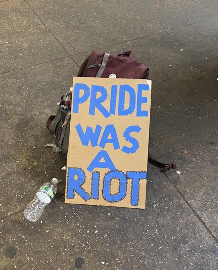 My latest handmade protest sign. Blue tape and black marker on rectangular cardboard. “pride was a riot” and riot is outlined in squiggly black. It leans against my burgundy and grey backpack, empty water bottle sitting on subway platform. The white pin on my bag is a tombstone reading “gender roles are dead”.