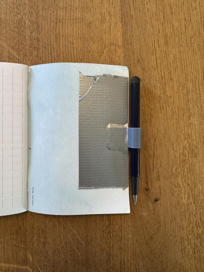 Back of a A6 notebook. There is a blue pen on the right side of the book. It’s slid into a greyish pen holder. The pen holder is glued to the page with a strip of duct tape. The book lays on a wooden table.