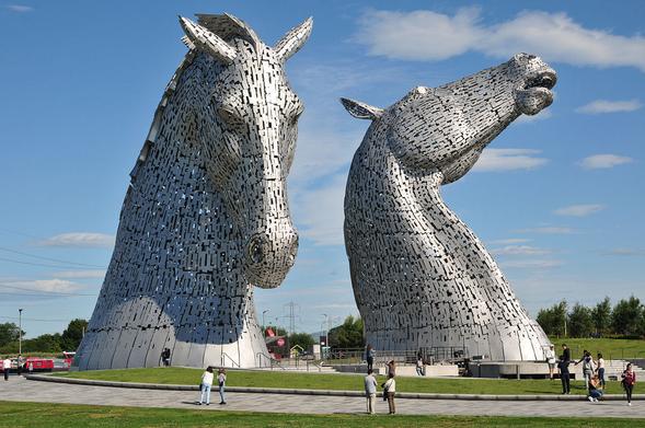 The Kelpies. The image shows the two tall sculptures of horses’ heads made from small plates of shiny silver metal. One is turned to face the camera, the other is looking up into the sky. In the foreground is a broad path on which some people are standing. The sky is largely blue.
