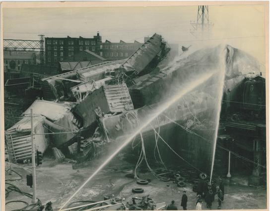 Firefighters spray water onto a huge jumble of wrecked train cars in an industrial city.