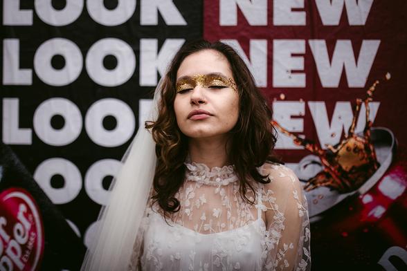 This is a bridal portrait, taken by Hamburg based European elopement and wedding photographer Sturmsucht, featuring a woman in an elegant white wedding dress with delicate lace detailing and a sheer overlay. She wears a flowing white veil and a striking golden masquerade mask decorated with glitter that sparkles across her eyes.

The bride has shoulder-length dark wavy hair and gazes upward with a serene expression. Her dress features intricate floral lace patterns and a high ruffled neckline.

Behind her is a bold graphic background with "LOOK" repeated in white text on black (left side) and "NEW" on red (right side). A coffee cup with a spider decoration appears in the lower right corner.

The portrait combines traditional bridal elegance with theatrical elements, creating an artistic and unconventional wedding photograph.