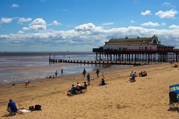 A busy beach scene on a sunny day in Cleethorpes with dramatic white clouds scattered across a bright blue sky. In the center, a Victorian pier extends into calm waters, featuring a traditional building with green roof and ornate ironwork supports. The golden sand beach is dotted with people enjoying the day - families in beach chairs, children playing, and groups sitting on towels. Some beachgoers wade in the shallow water near old wooden groynes that stretch into the sea. The wide beach stretches from foreground to the pier, creating a classic British seaside atmosphere with peaceful holiday activities throughout the scene.