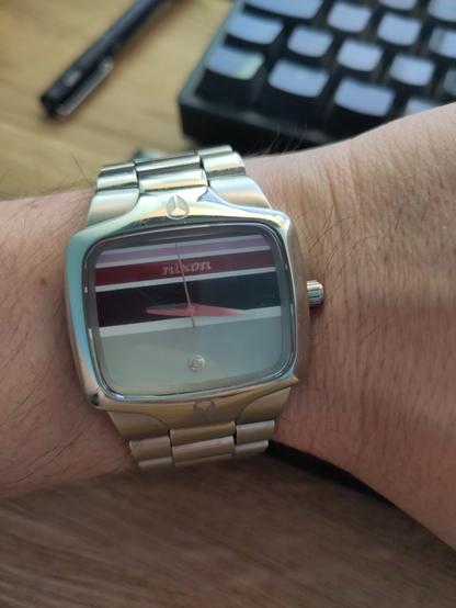 Photos of an analogue watch with squircle face, no numbers, and a metal bracelet strap. The face has a horizontal line pattern of burgundy, dark blue, and light blue. Nixon is written on the face and there is a tiny diamond.