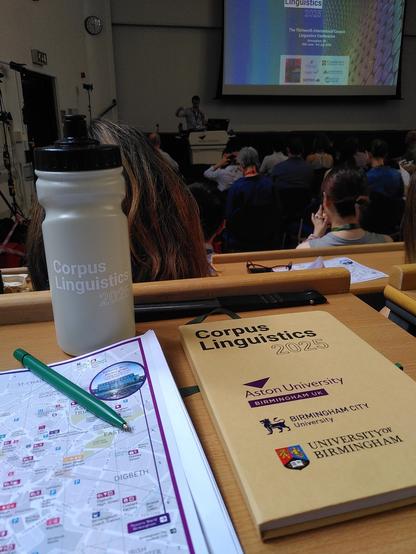 A conference table witha map, a water bottle and a notebook with conference logotype. In the background, a presenter and a large screen with a presentation of the Corpus Linguistics 2025.