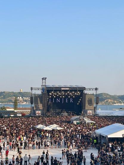 A large crowd is gathered at an outdoor music festival in front of a stage displaying the name "JINJER." The scene features a clear blue sky, and various tents and structures are visible in the foreground. The audience is predominantly wearing black clothing