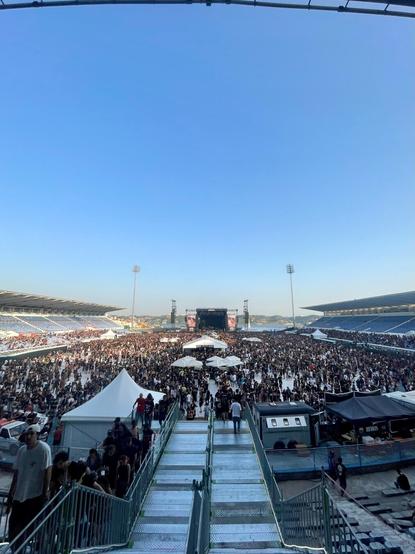 A large outdoor concert or festival scene with a massive crowd gathered in front of a stage. The crowd is densely packed, with various tents and structures visible. The background features a clear blue sky and stadium seating.