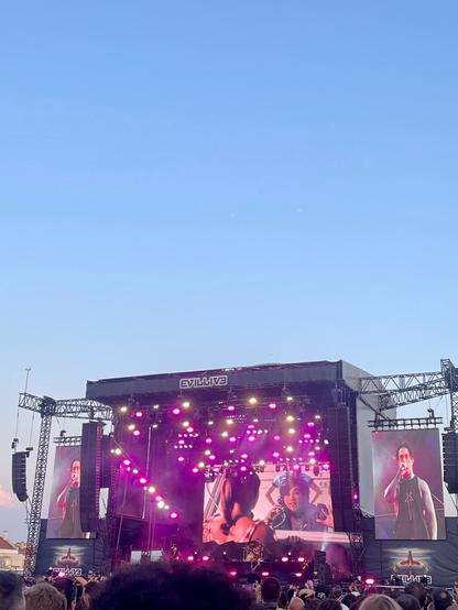 The image shows a live concert stage with vibrant purple lighting. Large screens display images of performers while an audience gathers in front. The sky is clear and blue in the background.