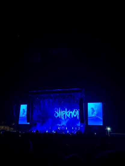 A concert scene featuring the band Slipknot on stage, lit by blue and purple lights. Large screens display a close-up of a band member. The audience is visible in silhouette.