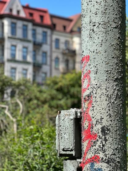 a utility pole and some fixtures, covered with lumpy grey paint and graffiti. In the background, one can see blurred greenery and apartment buildings.