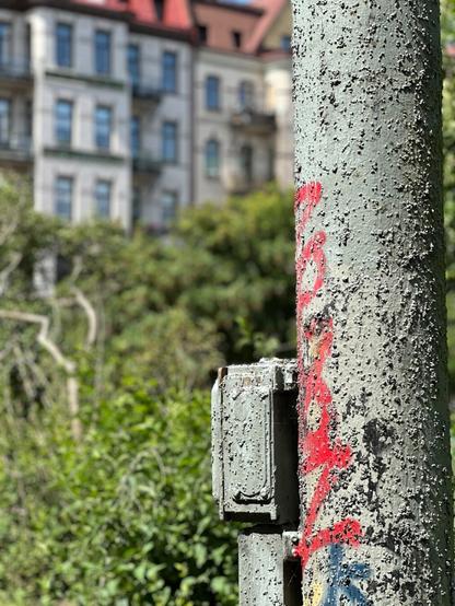 a utility pole and some fixtures, covered with lumpy grey paint and graffiti. In the background, one can see blurred greenery and apartment buildings.