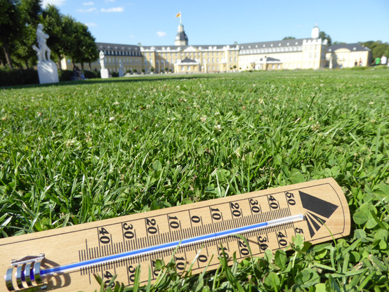 Symbolbild "Hohe Temperaturen am Schloss":
Das Bild zeigt eine sonnenbeschienene Rasenfläche mit einem darauf liegenden Thermometer aus Holz. Die Temperatur ist bei 35°. Im Hintergrund ist unscharf das karlsruher Schloss zu sehen.
© Umwelt- und Arbeitsschutz