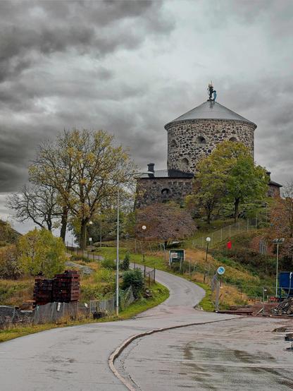 Vi ser Skansen Lejonet i Göteborg i bakgrunden, och en väg som sniglar sig från vårt perspektiv upp mot fortifikationen. Himlen är grå och trist, och asfalten är våt av regn.