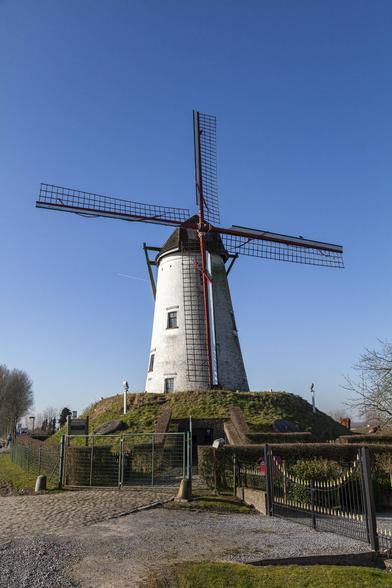 Crisp, clear blue sky forms the backdrop to a small rise of land just behind a gravel area with some wire fencing. On top of the low mound stands a nineteenth century windmill. It's a slightly tapering cylinder of thick stone painted white and on the left side which has most of the sunlight on it there are two windows offset from each other and a small, closed door in the base. The windmill has the trellis-like framework of four sails with two rising straight up and down, and two to either side at right angles.
