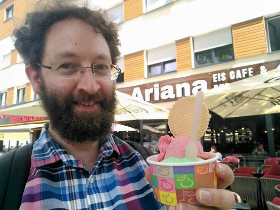 Manuel steht vor einer Eisdiele mit Eisbecher in der Hand: Waldmeister und Himbeere.