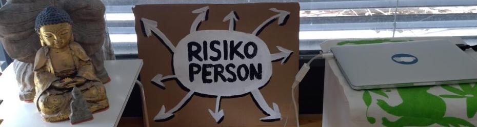 A sign reads "Risiko Person", which means person at risk, surrounded by arrows pointing in all directions. Some buddha statues and a laptop are visible