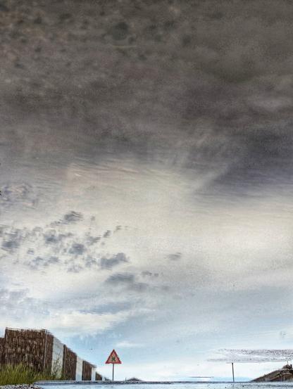 A vertical, wide shot captures a reflection of a cloudy sky and landscape in a puddle. The image is dominated by the grey and white tones of the sky, reflected with some distortion, showing ripples on the water's surface. In the lower third of the reflection, a triangular red and white warning sign with a black symbol of an X for crossroads is visible. To the left of the sign, there's a reflection of what appears to be a building or fence with a textured, brownish surface, and a patch of green grass is reflected in the very bottom left corner. To the right of the sign, a vertical line, possibly a pole, is reflected. The reflection on the right side of the puddle shows more indistinct shapes that could be distant buildings or structures. The overall impression is that of an ordinary scene transformed by its watery reflection.