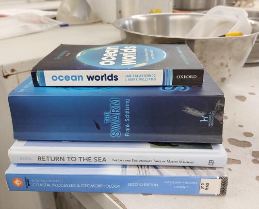 A stack of 4 blue books on my lab desk: Ocean Worlds by Jan Zakasiewicz & Mark Williams. The Swarm by Frank Schätzing, Return to the Sea by Annalisa Berta. Introduction to Coastal Processes & Geomorphology by Masselink, Hughes, & Knight.