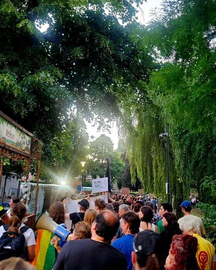 Een lange stoet demonstranten loopt door een groene parkachtige omgeving. Op de voorgrond is een regenboogvlag zichtbaar en iemand draagt een megafoon. Mensen houden protestborden omhoog.