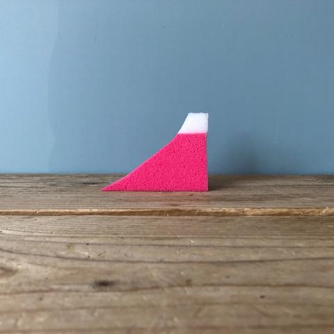 A small broken off segment of a pink sponge with a white top resting on a course wooden table up against a plain light blue wall.