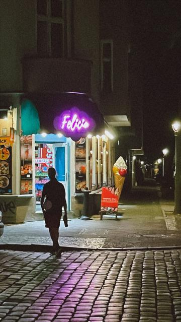Straße mit Kopfsteinpflaster bei Nacht, hellerleuchteter Eisladen an der Straßenecke, davor eine Silhouette