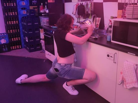 a picture of me squatting in front of a sink, draining pasta. I'm a white enby with a brown sidecut. I'm wearing denim shorts, white trainers and a black croptop. I'm squatting in front of a sink, draining pasta. my right leg is bent, my left leg almost fully extended and to my side. I'm holding the pot in front of me roughly on eye level just above the sink. there's an oven to my left and a microwave to my right, in the background one can see crates of bottles. the entire picture has purple lighting