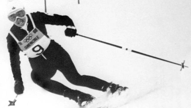 A black and white photograph depicts Nancy Greene in motion while downhill skiing at the Grenoble Olympics. She’s wearing a racing suit, goggles, and helmet, with bib number 9 clearly visible. The background shows a snow-covered slope.
