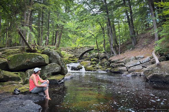 A person sits barefoot on a mossy boulder at the edge of a gently flowing forest stream, legs dangling as feet skim the water. The stream winds through dense trees and large rocks blanketed in green moss.