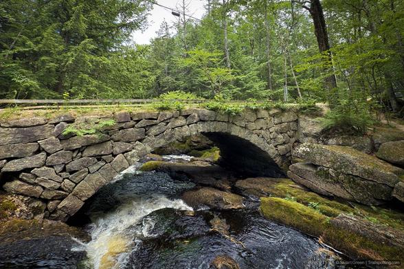 A rustic stone arch bridge spans a gently flowing forest stream, surrounded by lush greenery. The bridge, constructed from uneven, weathered stones, blends into the natural environment with moss clinging to its surface. Beneath it, clear water tumbles over mossy rocks, creating small cascades. Dense foliage and tall trees frame the scene.